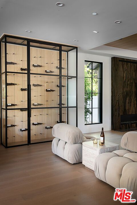 This interior shot showcases a modern living room with a prominent wine cellar display. The room features a neutral color palette, with a focus on natural light from a large window. The furniture includes a unique, modular sofa and a marble side table, contributing to a sophisticated and comfortable atmosphere.