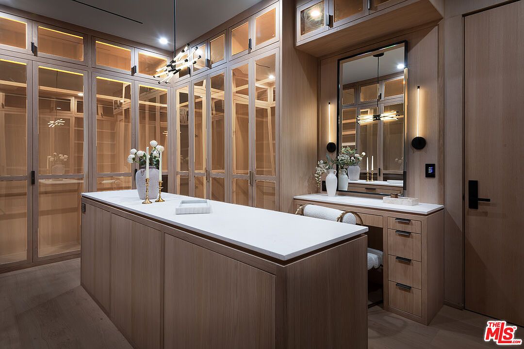 This is a well-organized walk-in closet featuring custom wood cabinetry with glass doors and a central island with a white countertop. The closet includes a vanity area with a mirror and decorative lighting, creating a luxurious and functional space. The overall design is elegant and maximizes storage.
