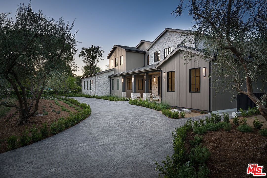 The image showcases the front exterior of a modern farmhouse-style home. A long, winding driveway made of gray pavers leads to the house, which features a combination of gray siding and stone accents. The landscaping includes manicured shrubs and trees, creating a welcoming and elegant curb appeal.