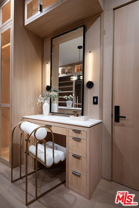 This is a well-lit primary bathroom featuring a vanity area with a large mirror and sconce lighting. The vanity has a white countertop and light wood cabinetry with sleek, dark hardware. A stylish chair with a gold frame and white cushions sits in front of the vanity, adding a touch of elegance.