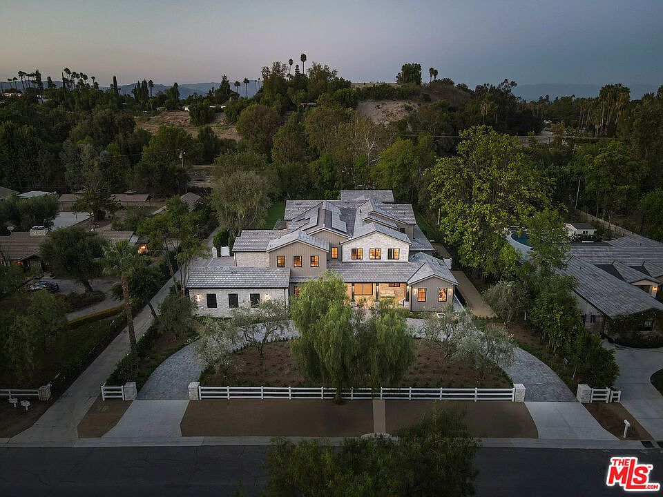 This aerial view showcases a sprawling, luxurious home with a meticulously landscaped yard. The house features a complex roofline, a mix of stone and siding, and numerous windows that suggest ample natural light. A white fence borders the property, adding to its curb appeal and sense of privacy.