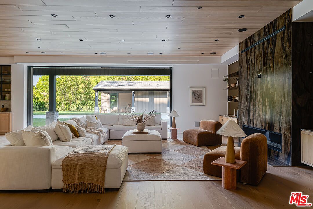 This is a bright and spacious living room featuring a large sectional sofa, a patterned area rug, and a modern fireplace with a dark wood surround. The room has a contemporary design with clean lines and a neutral color palette, complemented by natural light streaming in through a large sliding glass door. The open floor plan enhances the sense of space and luxury.