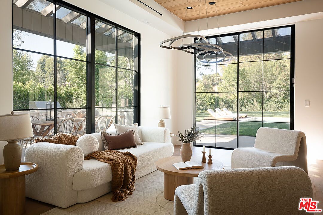 This is an interior shot of a modern living room featuring a plush, white sofa and matching armchairs arranged around a round coffee table. Large, black-framed windows offer a view of the outdoor greenery and pool area, enhancing the sense of spaciousness and connection to nature. A unique, multi-ring chandelier hangs above, adding a touch of contemporary elegance.
