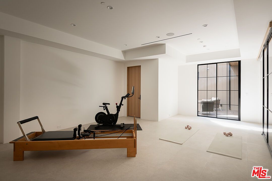 This is a bright and spacious home gym featuring a pilates reformer, stationary bike, and yoga mats with dumbbells. The room has white walls, recessed lighting, and large windows that provide natural light. The overall impression is clean, modern, and conducive to a healthy lifestyle.