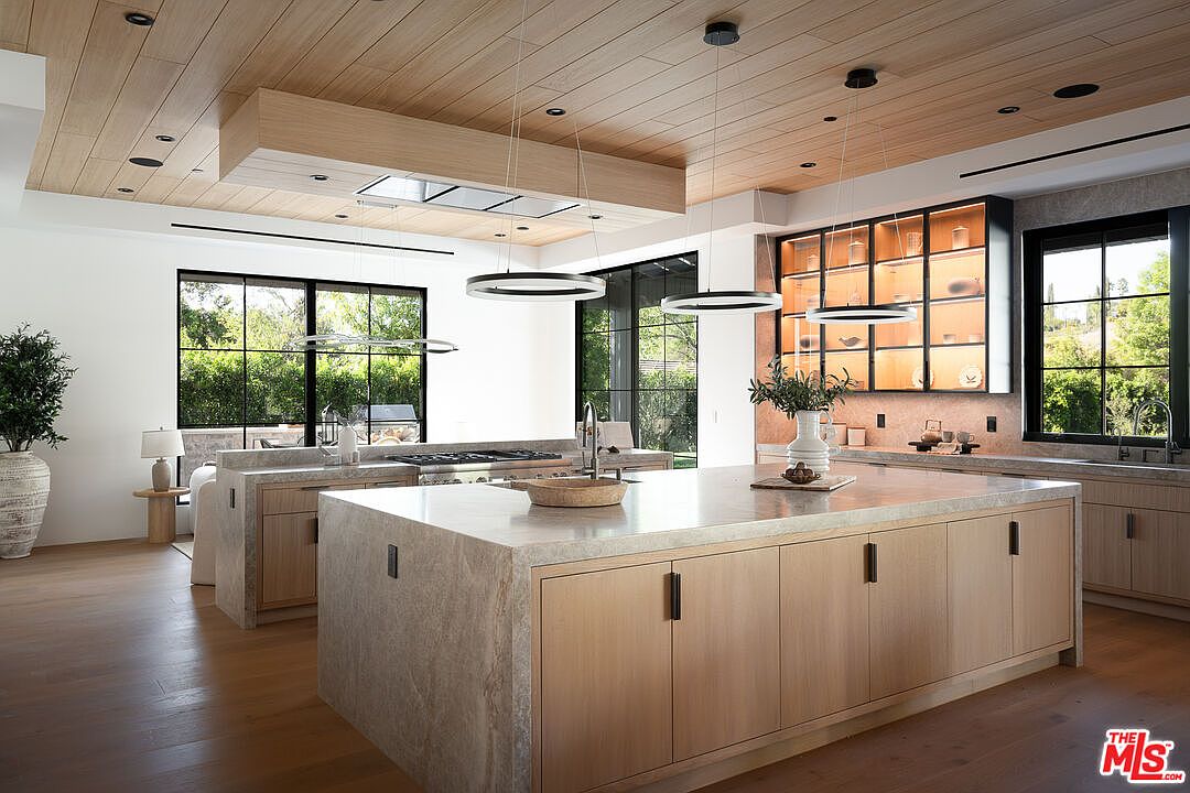 This is a bright and modern kitchen featuring a large island with a stone countertop and light wood cabinetry. The kitchen includes stainless steel appliances, pendant lighting, and large windows that offer views of the lush greenery outside. The overall design is sleek and contemporary, creating a welcoming and functional space.