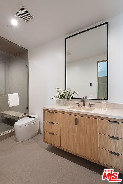 This is a modern primary bathroom featuring a floating vanity with light wood cabinetry and a light countertop. A large rectangular mirror hangs above the vanity, reflecting the spacious interior. A sleek, modern toilet sits next to a glass-enclosed shower with a built-in bench, creating a spa-like atmosphere.