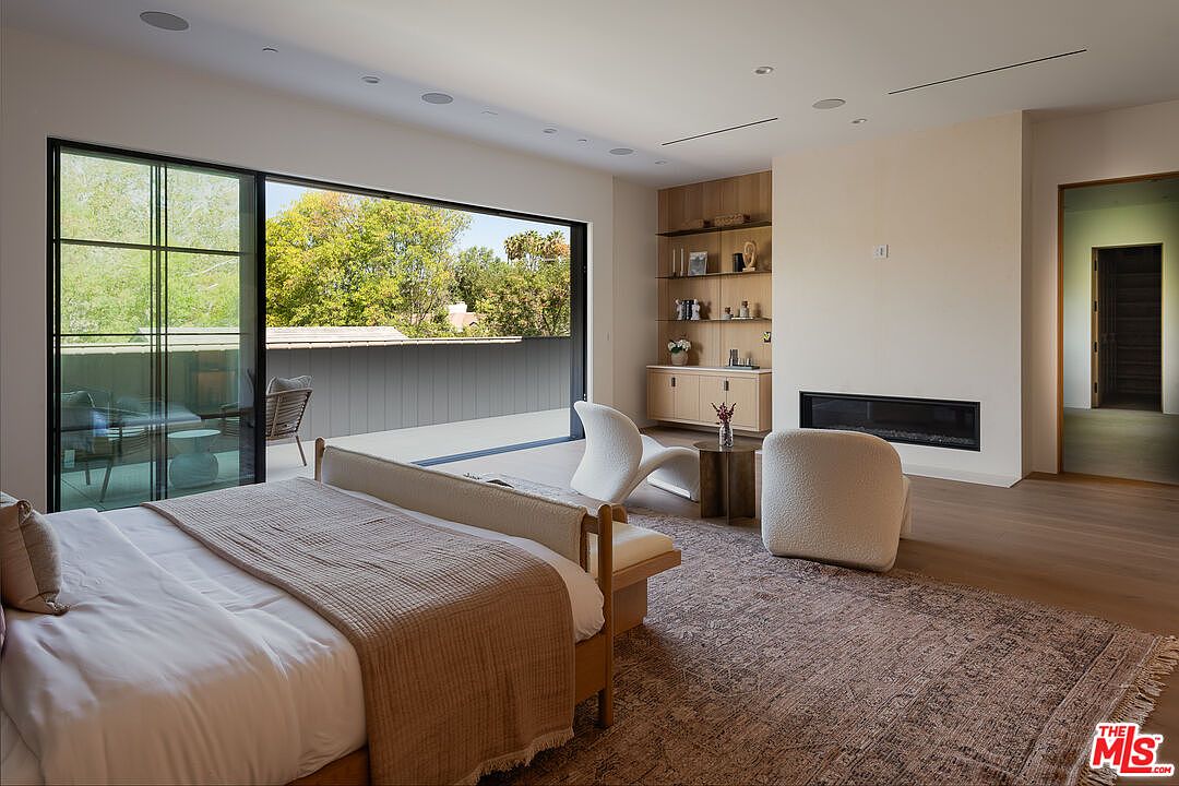 This is a primary bedroom featuring a large bed with a beige throw, a seating area with a modern white chair and a fluffy chair, and a built-in shelving unit with a fireplace. A large sliding glass door opens to a balcony, providing natural light and a view of the outdoors. The room is decorated in neutral tones with hardwood floors and a patterned rug, creating a serene and luxurious atmosphere.