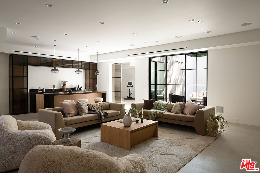 This is a spacious and modern living room featuring two beige sofas, two plush armchairs, and a large wooden coffee table on a patterned rug. The room has a contemporary design with a bar area in the background and large windows providing natural light. The overall impression is one of comfort and sophistication.