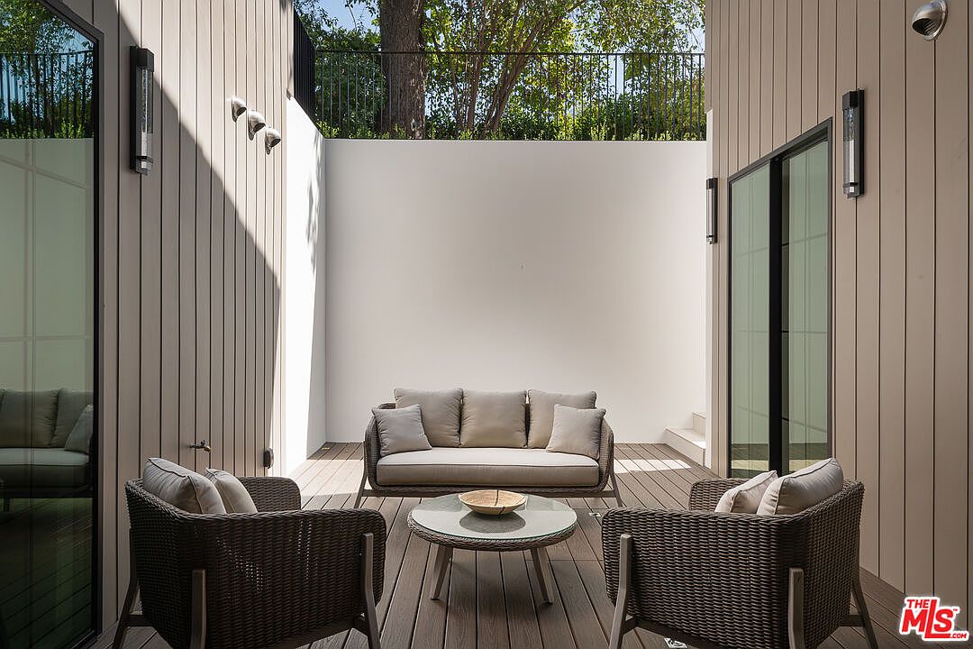 This image showcases a stylish outdoor patio area, featuring modern furniture including a sofa, two armchairs, and a round coffee table. The patio is constructed with wooden decking and is enclosed by walls, providing privacy. The space is well-lit with wall-mounted light fixtures, creating a cozy and inviting atmosphere, perfect for relaxation or entertaining.