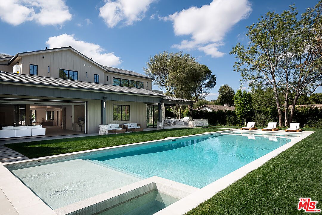 This image showcases a luxurious backyard featuring a rectangular pool with a built-in spa and shallow tanning ledge. The pool is surrounded by lush green grass and is adjacent to a modern home with large sliding glass doors that open to an outdoor living space. Lounge chairs are placed near the pool, creating an inviting and relaxing atmosphere.