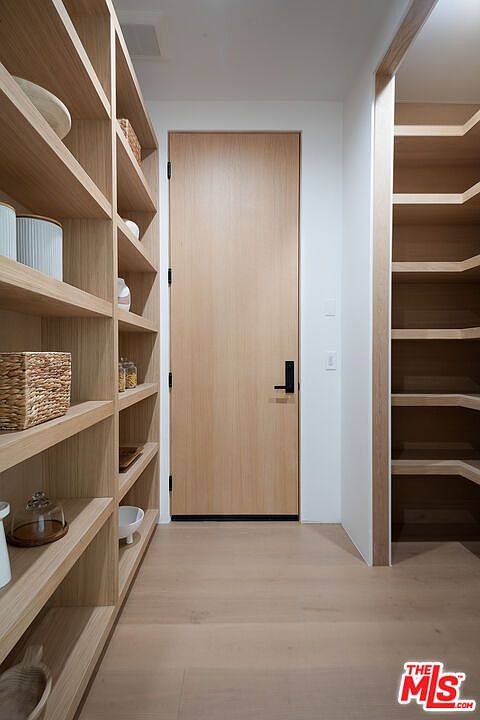 This is an interior shot of a well-organized pantry. The pantry features light wood shelving on both sides, with various containers and items neatly arranged. A light wood door with a black handle is visible at the end of the pantry, and the flooring is light wood as well, creating a clean and modern aesthetic.