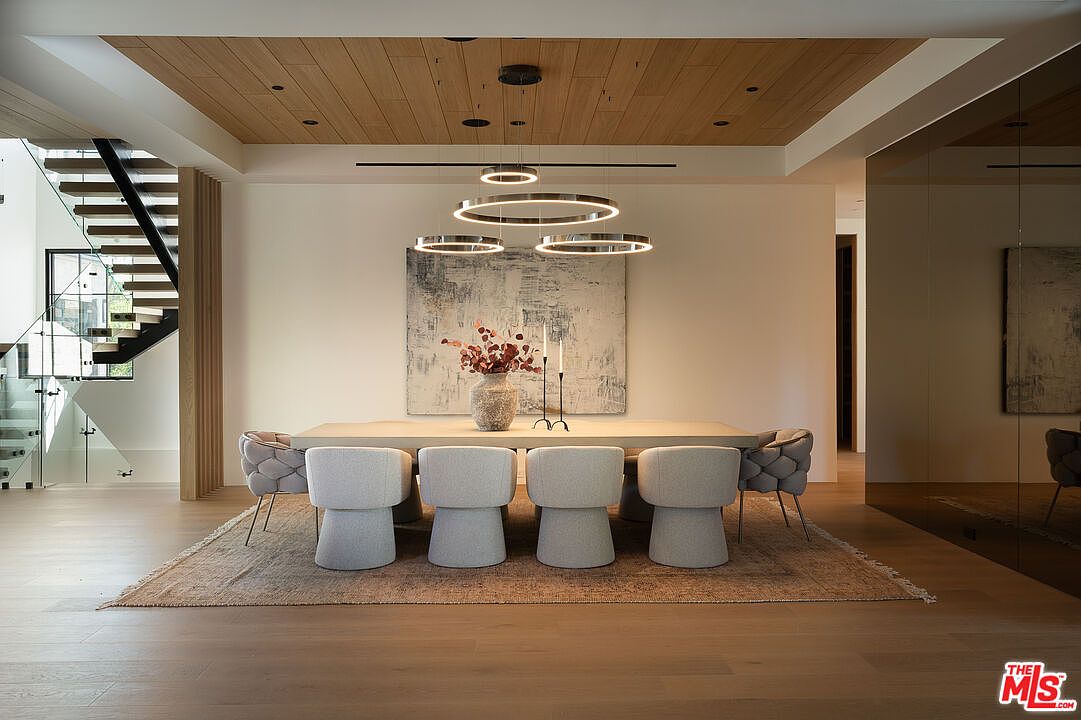 This is a dining room featuring a long, light-colored table with modern chairs arranged around it. A striking circular chandelier hangs above the table, and an abstract painting adorns the wall. The room is well-lit and has a contemporary design, with wooden floors and a wooden ceiling accent.