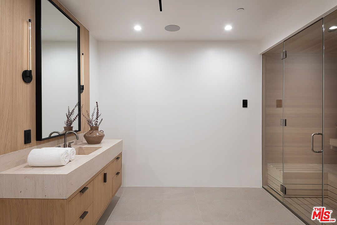 This is a modern primary bathroom featuring a light wood vanity with a stone countertop and integrated sink. A large mirror with black trim is mounted above the vanity, flanked by sconce lighting. A glass-enclosed sauna is visible on the right, adding a spa-like feel to the space. The overall aesthetic is clean, minimalist, and luxurious.