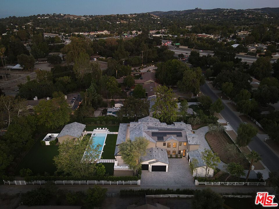 This aerial shot showcases a sprawling estate with a large main house, a detached garage, and a long rectangular swimming pool. The property is surrounded by lush greenery and mature trees, offering privacy and a sense of seclusion. A white fence encloses the property, adding to its charm and curb appeal.