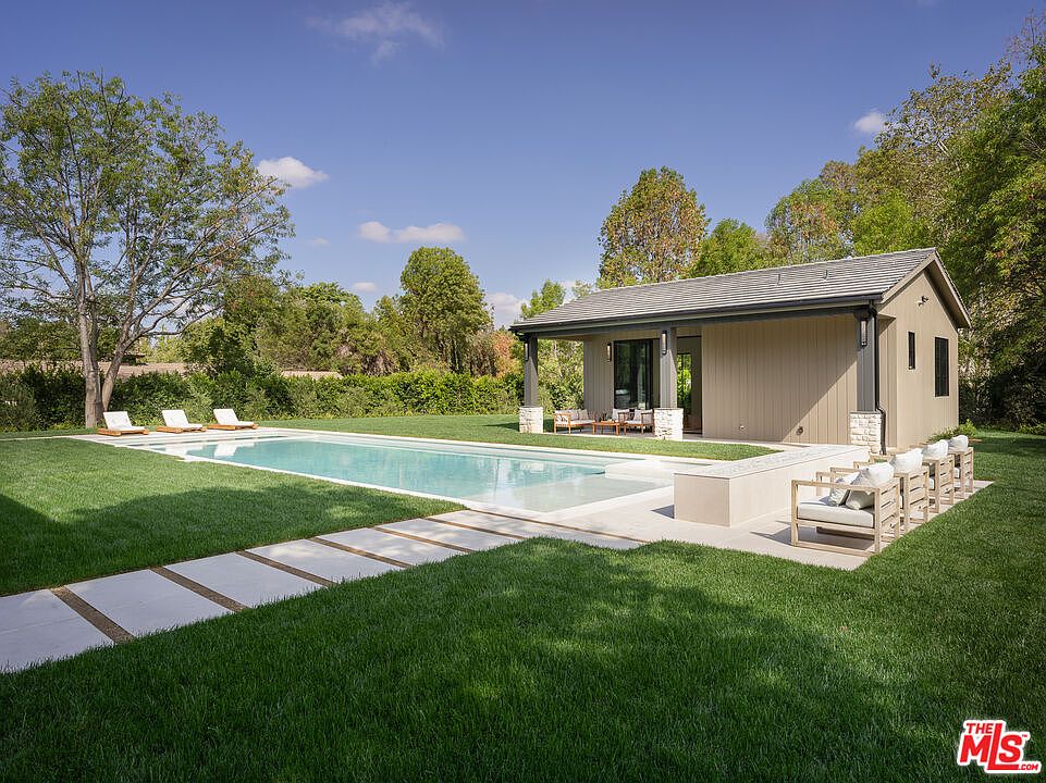 This exterior shot showcases a luxurious pool and spa area, complete with a modern pool house. The pool is surrounded by lush green grass and a stone walkway, leading to lounge chairs. The pool house features a covered patio with outdoor seating, creating an inviting space for relaxation and entertainment.