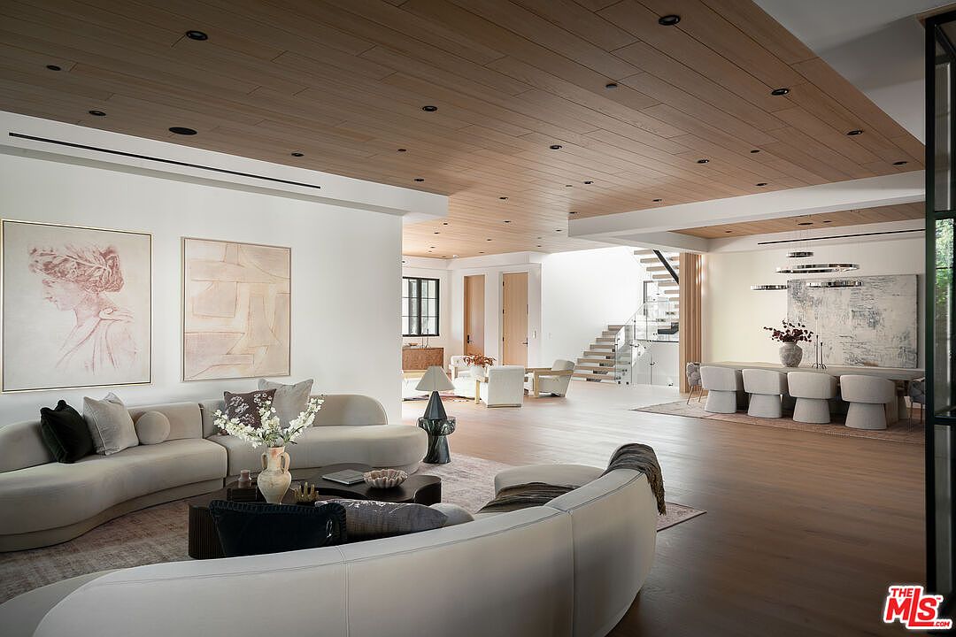 This is a spacious and luxurious living room featuring a curved sofa, modern artwork, and a wooden plank ceiling with recessed lighting. The open floor plan seamlessly connects the living area to the dining space and staircase, creating a bright and airy atmosphere. The hardwood flooring and neutral color palette contribute to the room's sophisticated and inviting ambiance.