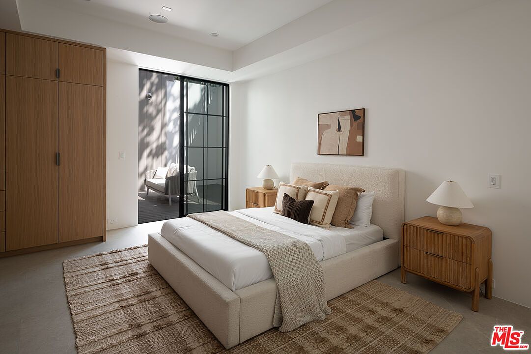 This is a bright and airy primary bedroom featuring a neutral color palette. The room includes a large bed with a textured headboard, two wooden nightstands with lamps, and a spacious wardrobe. A sliding glass door leads to an outdoor patio, enhancing the room's appeal and creating a seamless indoor-outdoor flow.