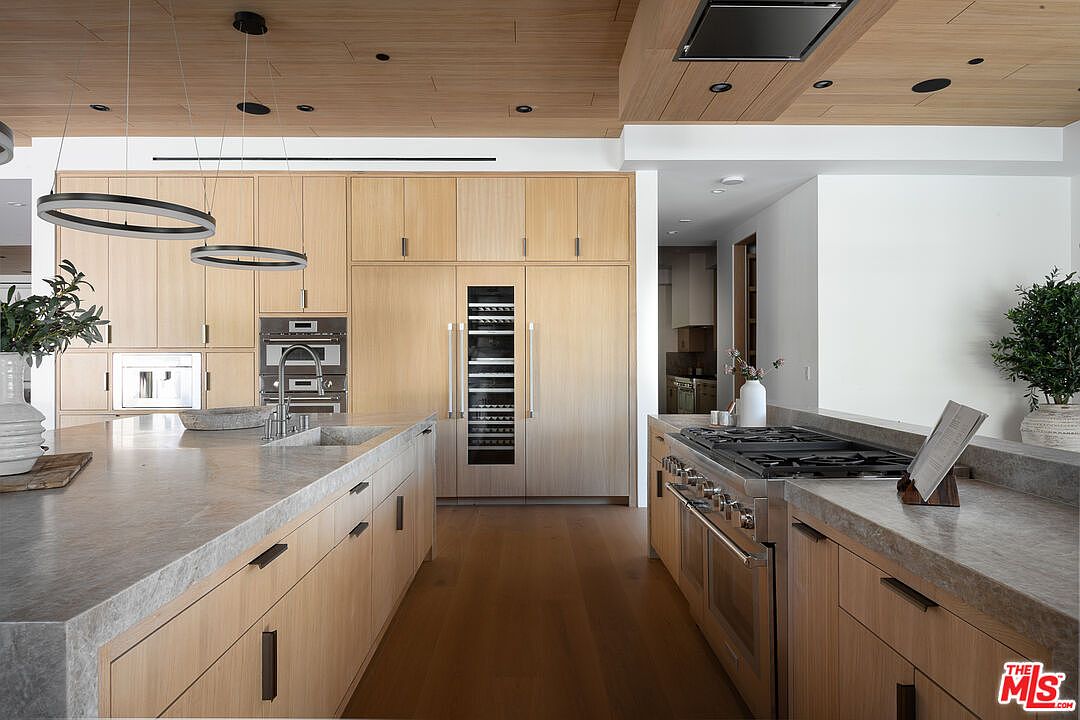 This is a modern kitchen featuring light wood cabinetry, stainless steel appliances, and a large island with a gray countertop. The kitchen includes a built-in wine fridge and a double oven. The ceiling is wood paneled with recessed lighting, and the overall style is sleek and contemporary.