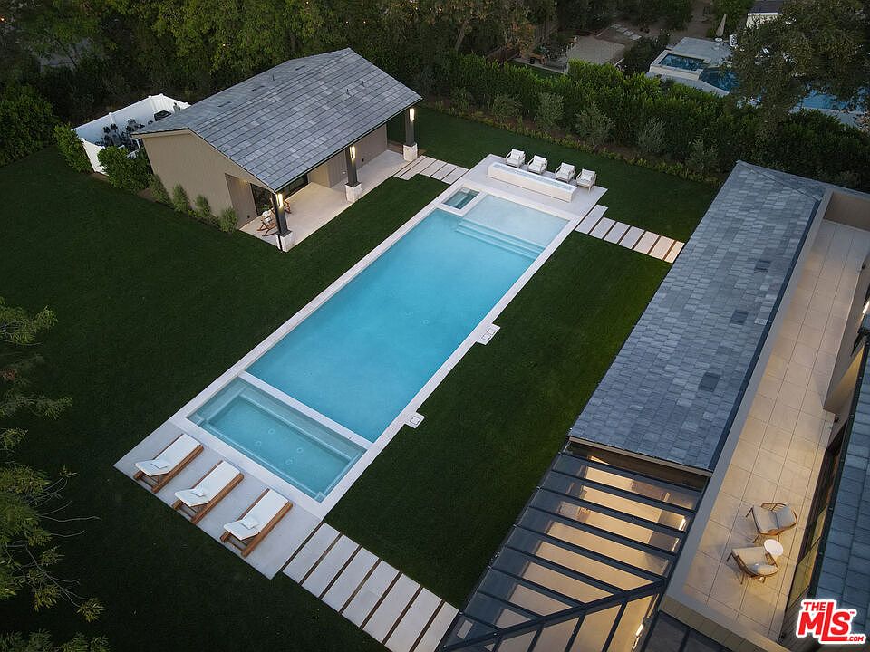 This aerial shot showcases a luxurious backyard featuring a long, rectangular pool with an integrated spa. The pool is surrounded by lush green grass and modern hardscaping, including stone pathways and lounge areas with deck chairs. A pool house and a portion of the main residence are also visible, highlighting the property's upscale design and outdoor living spaces.