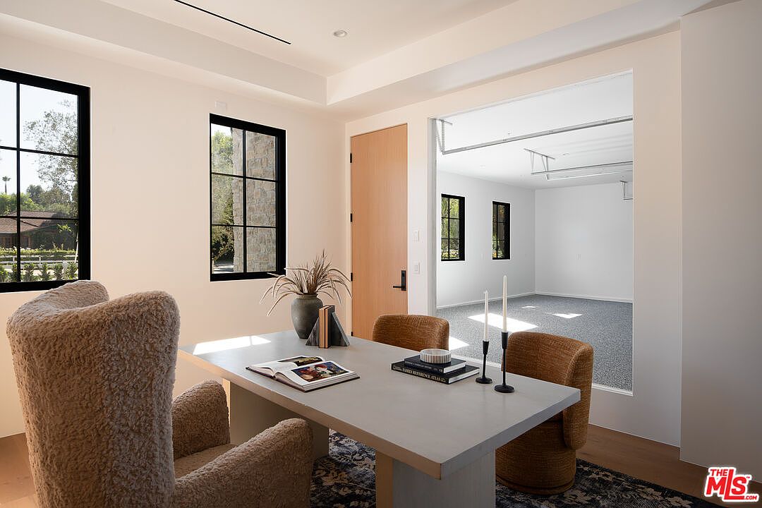This is an interior shot of a modern office space featuring a large desk with books and decorative items, comfortable chairs, and two windows providing natural light. The room has a neutral color palette with light walls and a dark rug, creating a sophisticated and inviting atmosphere. An open doorway leads to another room, adding depth and visual interest.
