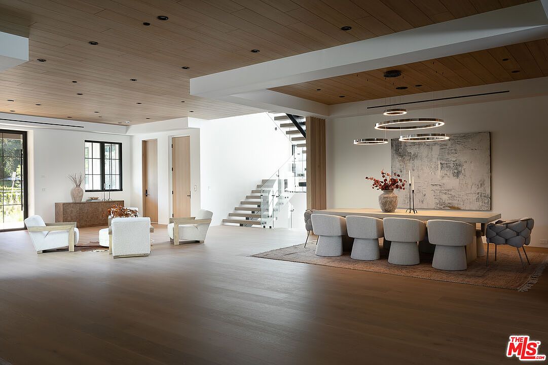 This is a spacious dining room featuring a long, light-colored wooden table surrounded by modern gray chairs. Above the table, there are circular pendant lights, and a large abstract painting hangs on the wall. The room has hardwood floors and a staircase in the background, creating an open and airy atmosphere.