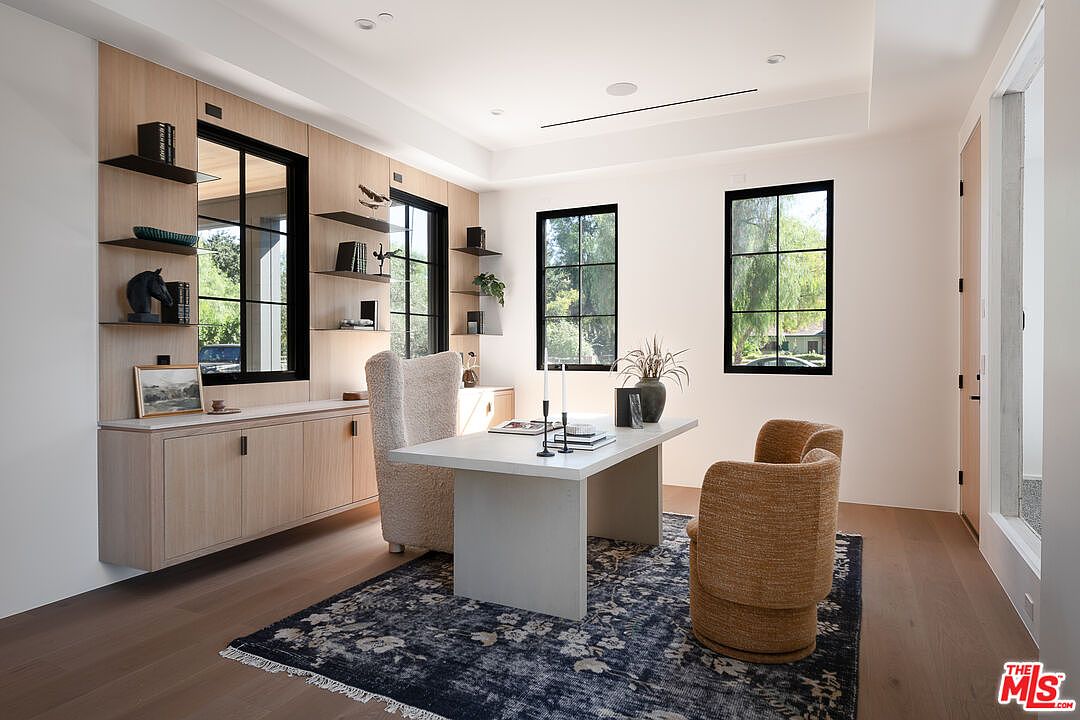 This is an interior shot of a modern home office featuring built-in shelving and cabinetry in a light wood finish. A large desk sits in the center of the room, flanked by two stylish chairs, and a dark blue patterned rug anchors the space. Natural light floods the room through several windows, creating a bright and inviting workspace.