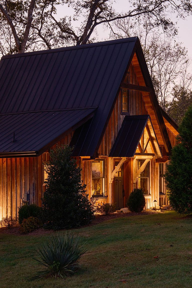 The image showcases the front exterior of a charming, rustic cabin-style home. The house features a dark metal roof, warm wood siding, and inviting lighting that highlights the architectural details. The well-maintained lawn and landscaping add to the property's curb appeal, creating a welcoming and picturesque scene.