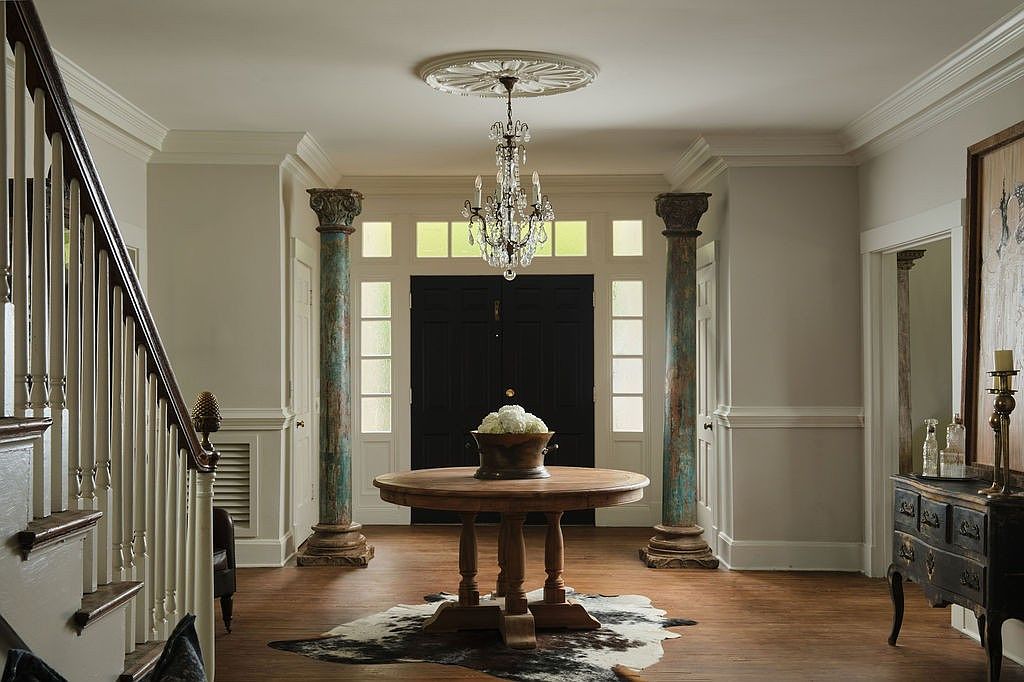 This is an interior shot of a grand entryway featuring hardwood floors, decorative columns, and a striking chandelier. A round wooden table with a floral arrangement sits atop a cowhide rug, adding a touch of rustic elegance. The staircase on the left and a glimpse of a console table on the right suggest a spacious and well-appointed home.