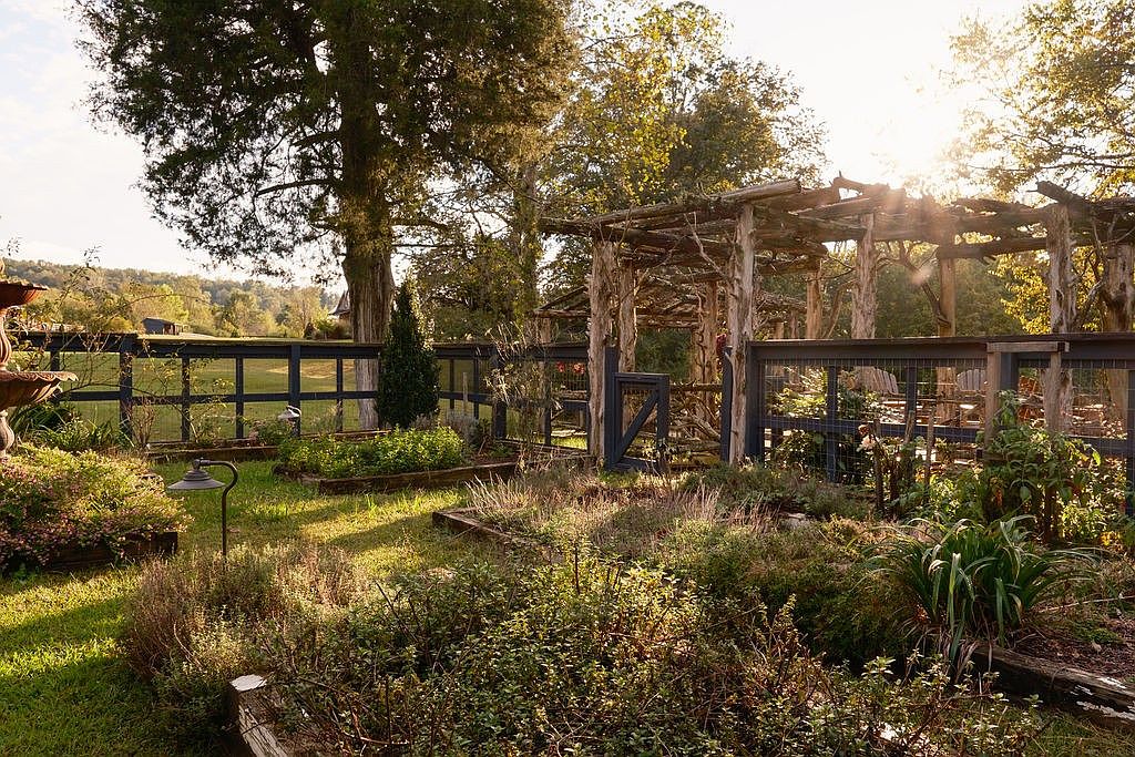 This image showcases a charming garden with a rustic wooden pergola and a dark-colored fence. The garden beds are filled with lush greenery, creating a serene and inviting atmosphere. Sunlight filters through the trees, adding warmth and highlighting the natural beauty of the outdoor space.