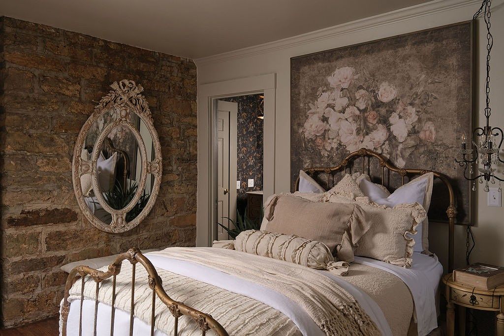 This is a bedroom featuring a bed with a gold frame, a stone wall with an ornate mirror, and a floral painting above the bed. The room has a vintage, romantic style with soft lighting and neutral colors. A doorway leads to another room with patterned wallpaper, adding depth and interest.
