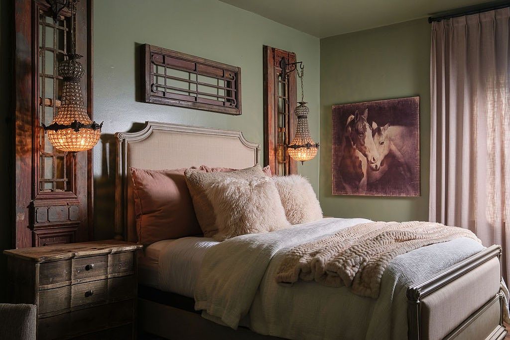 This is a cozy primary bedroom featuring a neutral-toned upholstered bed with plush pillows and a soft throw blanket. The room is decorated with rustic wooden accents, including a nightstand and decorative wall pieces. Two ornate chandeliers provide warm lighting, enhancing the room's inviting and comfortable atmosphere.