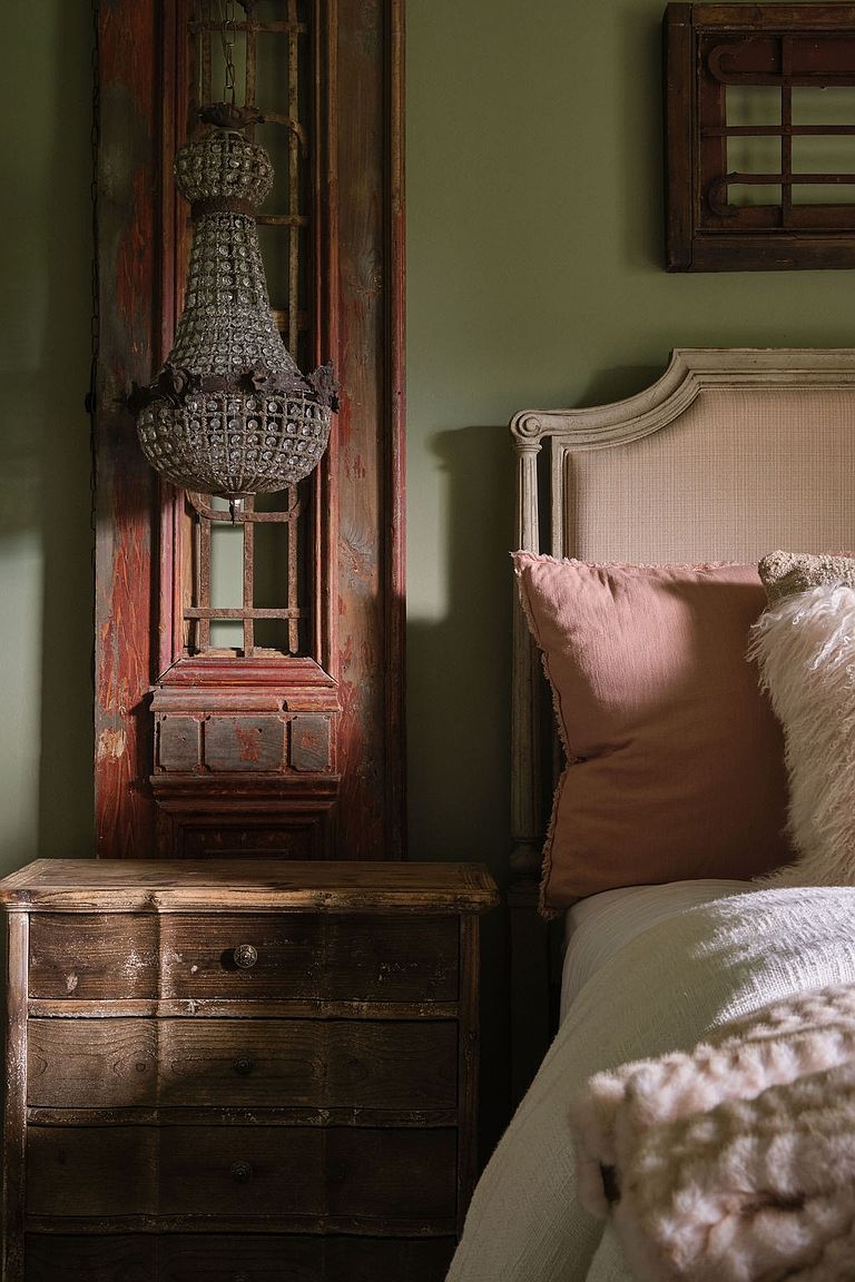 This image showcases a bedroom with a rustic and vintage aesthetic. A wooden nightstand sits beside a bed with a neutral-toned headboard and soft bedding, complemented by decorative pillows. An antique wooden door with a chandelier hanging in front of it serves as a unique wall decoration, adding character to the space. The room is painted in a muted green tone, enhancing the cozy and inviting atmosphere.