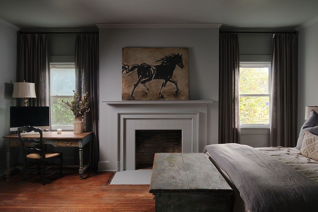 This primary bedroom features a decorative fireplace with a horse painting above the mantel, flanked by windows with dark curtains. A wooden desk sits to the left, while a bed with neutral bedding and a wooden chest at its foot occupies the right side of the frame. The room has hardwood floors and a calming, neutral color palette, creating a cozy and sophisticated atmosphere.