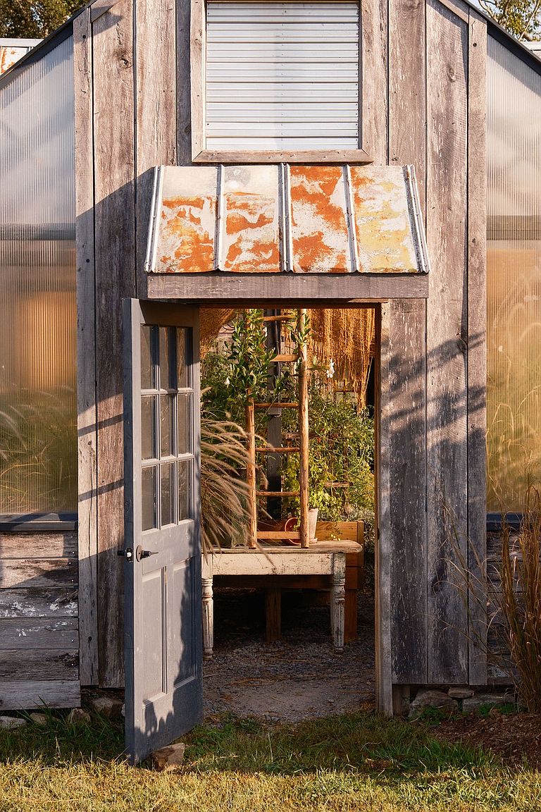 This image showcases a charming greenhouse with an open door, revealing a glimpse of the interior filled with plants and a rustic ladder. The weathered wood exterior and the surrounding greenery create a serene and inviting atmosphere, perfect for a garden enthusiast. The natural light filtering through the structure adds warmth and highlights the potential for cultivating a thriving garden space.