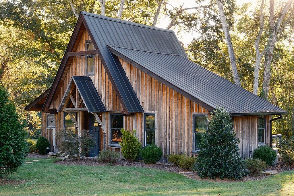 The image showcases the front exterior of a charming, rustic house with a unique architectural design. The house features wooden siding, a dark metal roof, and a prominent gabled entrance. Lush greenery surrounds the property, enhancing its curb appeal and creating a serene, natural setting.