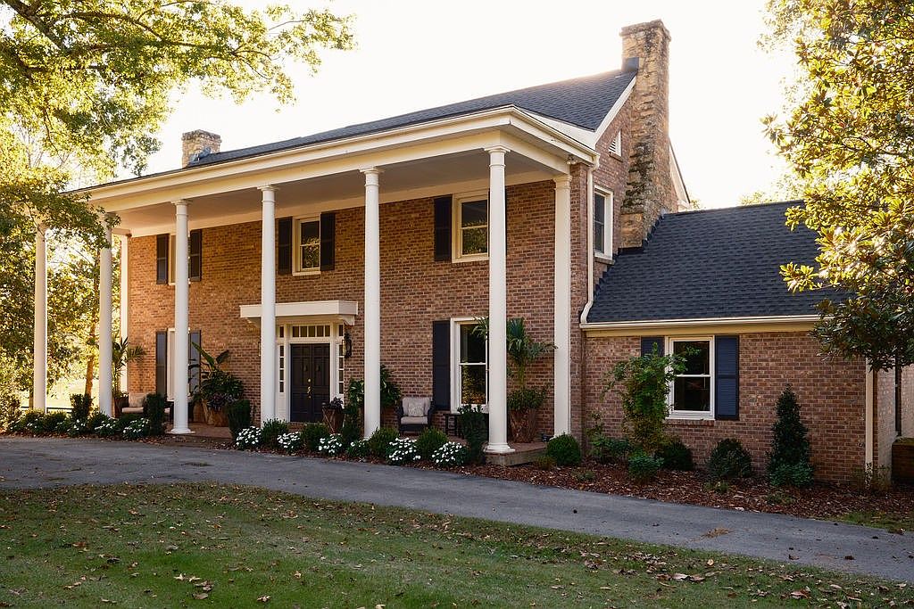 This is a front view of a stately brick home with a prominent, columned front porch. The house features a dark roof, dark shutters, and a well-manicured lawn, creating a classic and inviting curb appeal. A paved driveway leads up to the house, flanked by mature trees and landscaping.