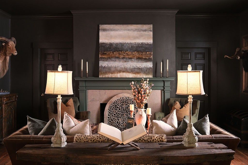 This is an interior shot of a living room featuring dark, moody walls and elegant decor. The focal point is a wooden coffee table with an open book, flanked by lamps and decorative objects. The room includes a fireplace with an abstract painting above it, and comfortable seating with decorative pillows, creating a sophisticated and inviting atmosphere.