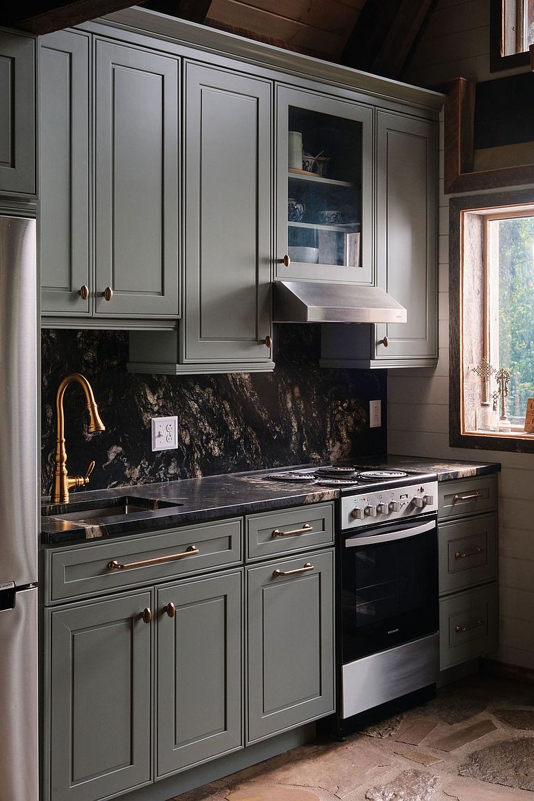 This is a cozy kitchen featuring gray cabinetry with bronze hardware, a black marble backsplash and countertops, and stainless steel appliances. A gold faucet adds a touch of elegance, while a window provides natural light. The overall impression is a blend of rustic charm and modern sophistication.