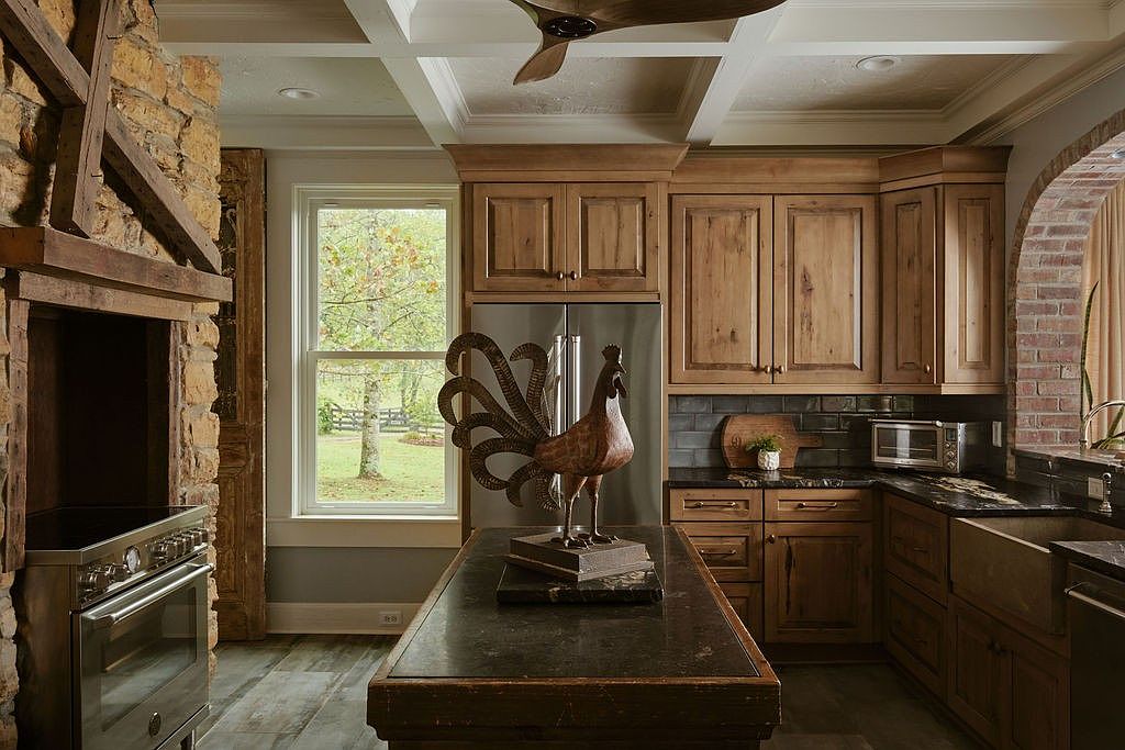 This is a warm and inviting kitchen featuring custom wood cabinetry, granite countertops, and stainless steel appliances. A large island with a dark countertop and decorative rooster statue sits in the center of the room. The kitchen also features a stone fireplace and a window overlooking a green yard, creating a cozy and rustic atmosphere.
