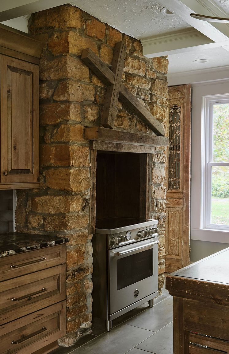 This image showcases a rustic kitchen interior, featuring a stone oven surround with wooden beams and a stainless steel range. The kitchen also includes wooden cabinetry with drawers and a countertop with dark stone. The overall impression is one of warmth and character, blending modern appliances with traditional design elements.