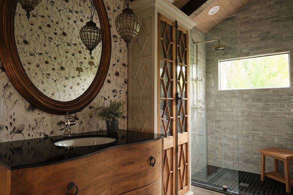 This is a stylish primary bathroom featuring a unique vanity with a dark countertop and an oval mirror. The walls are adorned with floral wallpaper, complemented by decorative pendant lights. A glass-enclosed shower with gray brick tiles and black hexagonal floor tiles adds a modern touch, while a wooden stool provides seating.