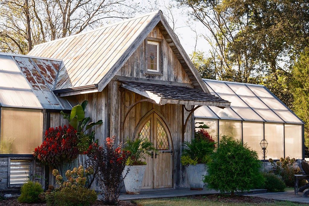 The image showcases the front view of a charming greenhouse structure with a rustic, cottage-like aesthetic. The building features weathered wooden siding, a metal roof, and arched double doors, complemented by lush greenery and potted plants. The overall impression is one of quaint elegance and natural beauty, making it an appealing feature for a unique property.
