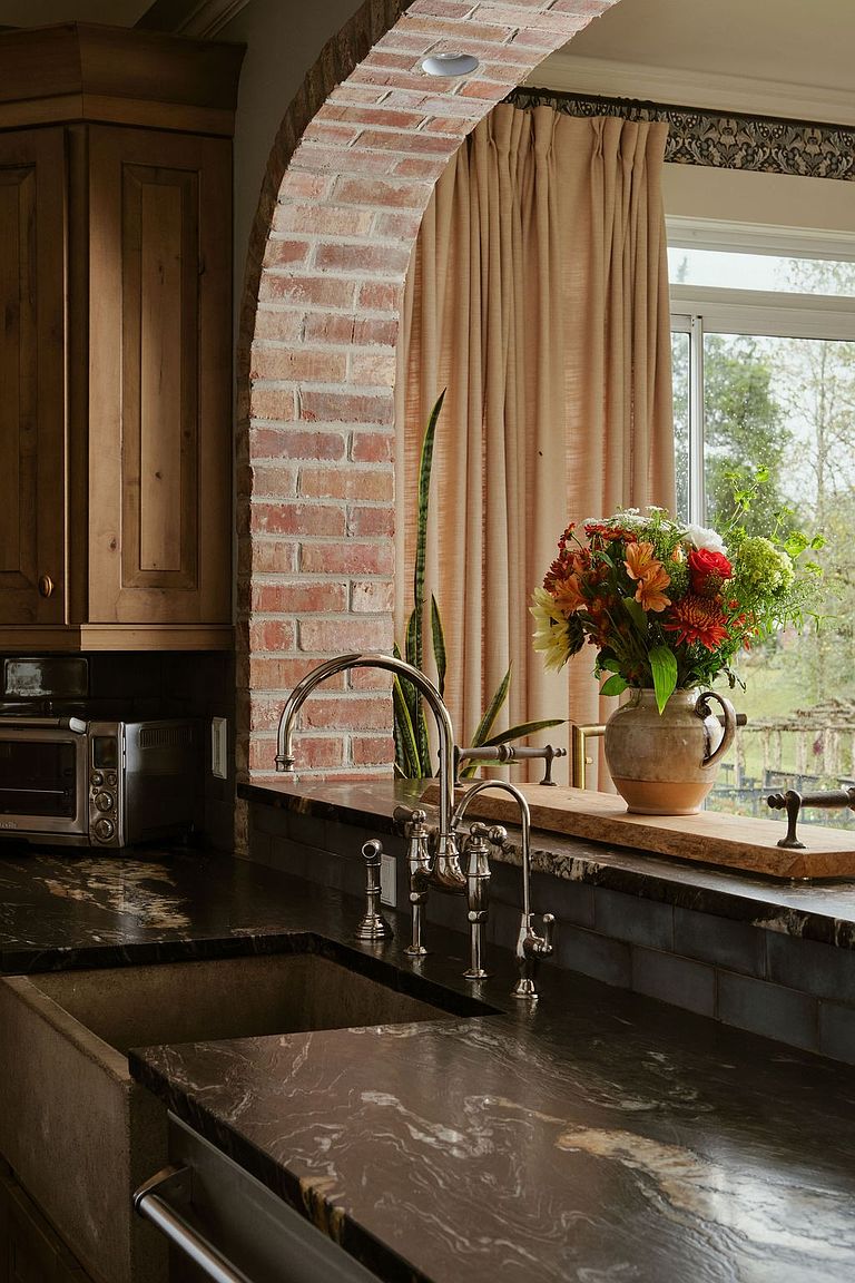 This kitchen features a unique brick archway, dark countertops, and a farmhouse sink, blending rustic charm with modern elegance. A bouquet of flowers sits by the window, adding a touch of nature to the space. The stainless steel faucet and appliances complement the dark cabinetry, creating a warm and inviting atmosphere.