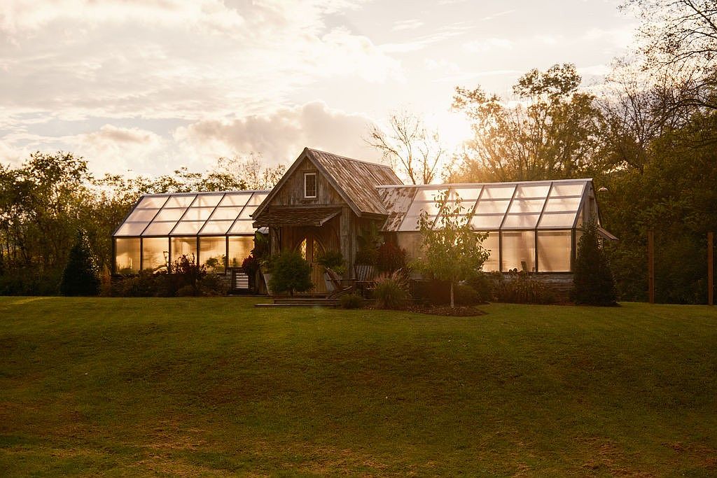 This image showcases a charming greenhouse connected to a rustic wooden structure, set against a backdrop of lush greenery and a golden sunset. The well-maintained lawn in the foreground enhances the property's appeal, creating a serene and inviting atmosphere. The architectural style blends seamlessly with the natural surroundings, suggesting a peaceful retreat.