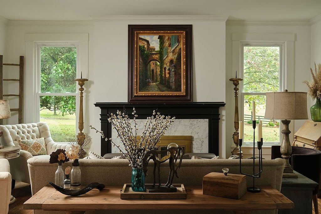 This is an interior shot of a living room featuring a neutral color palette with white walls and a beige sofa. A dark wood fireplace mantel is adorned with a framed painting, and the room is decorated with various decorative items such as candlesticks, vases, and a wooden box. Natural light streams in through the windows, creating a warm and inviting atmosphere.