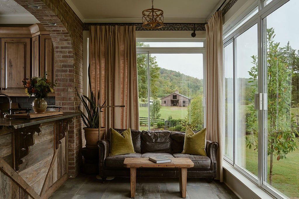 This sunroom features a comfortable leather sofa with olive green pillows, a rustic wooden coffee table, and large windows offering a view of a barn in the distance. The room is decorated with neutral-toned curtains and a unique light fixture, creating a cozy and inviting atmosphere. The combination of natural light and rustic elements makes this a desirable space for relaxation.