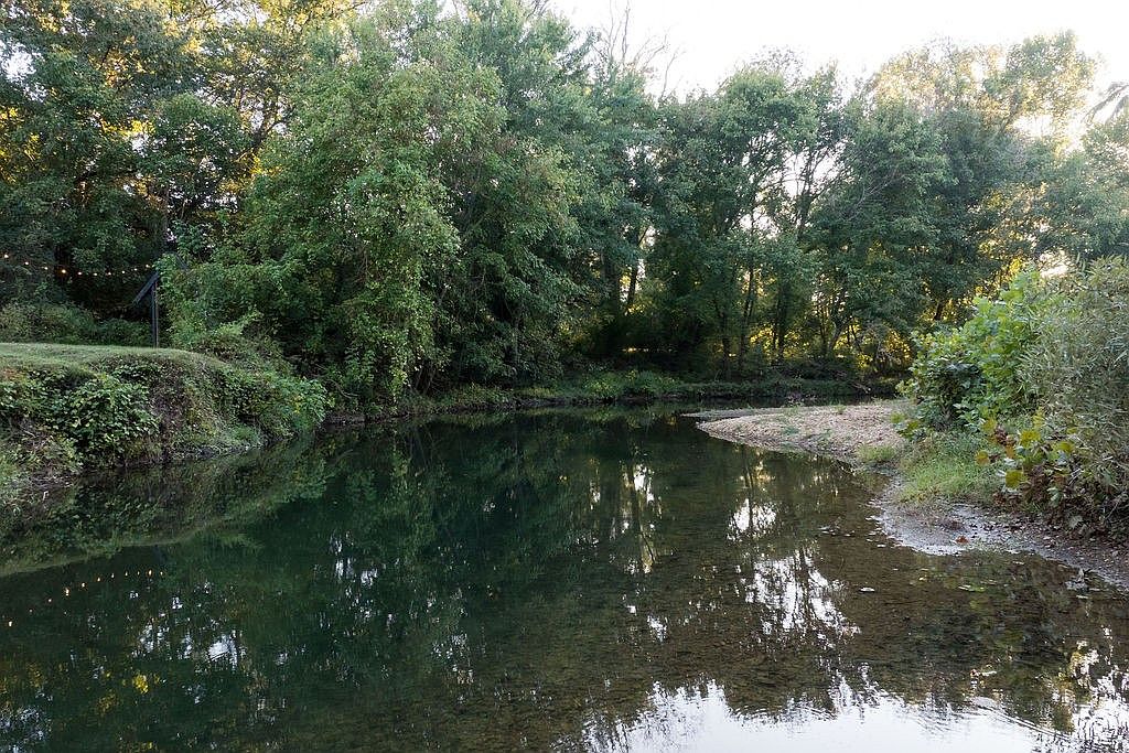 This serene outdoor scene showcases a tranquil river or creek bordered by lush greenery and mature trees. The water reflects the surrounding foliage, creating a peaceful and inviting atmosphere. A small sandy bank is visible on one side, suggesting a natural and accessible waterfront property.