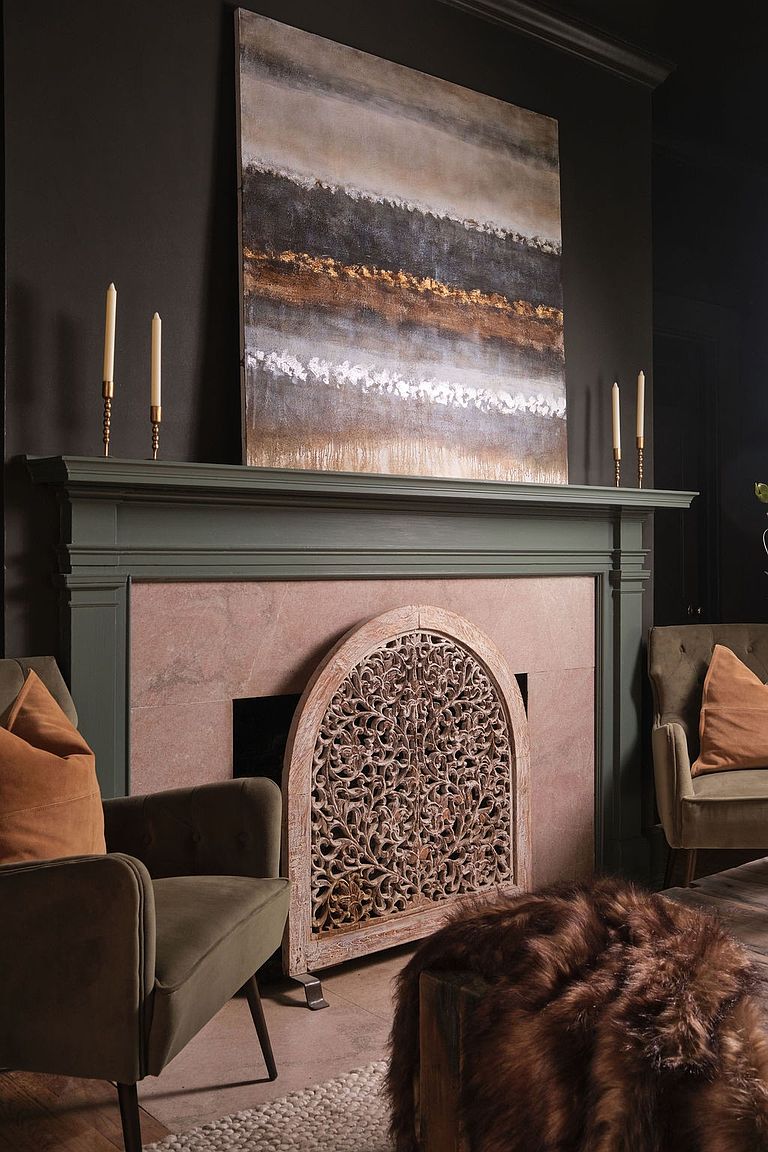 This image showcases a sophisticated living room interior, featuring a dark-painted wall, a fireplace with a decorative screen, and an abstract painting above the mantel. Two olive-green armchairs with orange pillows flank the fireplace, adding a touch of color and comfort. The room exudes a cozy and elegant atmosphere, perfect for relaxation and entertaining.