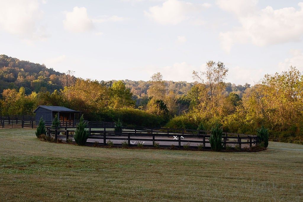 The image showcases a well-maintained yard with a fenced riding arena. A small barn-like structure is visible in the background, nestled among lush trees and foliage. The scene evokes a sense of tranquility and rural charm, ideal for equestrian enthusiasts or those seeking a peaceful country estate.
