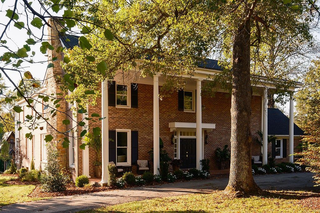The image showcases the front facade of a stately brick home with prominent white columns supporting a covered porch. Dark shutters accent the windows, and mature trees frame the house, adding to its curb appeal. The well-manicured lawn and landscaping enhance the property's inviting atmosphere.